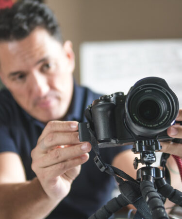 Homem jovem, profissional do audiovisual, realizando a captação de imagens com câmeras e microfone em local a céu aberto.