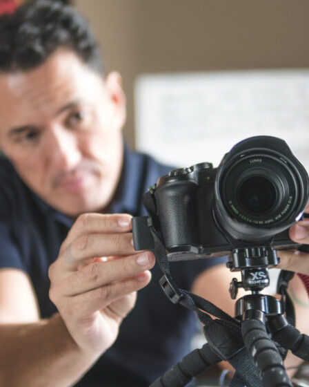 Homem jovem, profissional do audiovisual, realizando a captação de imagens com câmeras e microfone em local a céu aberto.
