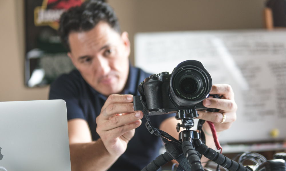Homem jovem, profissional do audiovisual, realizando a captação de imagens com câmeras e microfone em local a céu aberto.