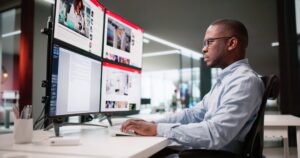 Jornalista em escritório trabalhando analisando monitores de computador.