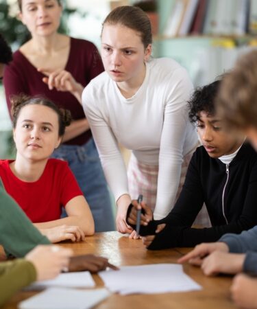 Alunos envolvidos discutindo e realizando uma atividade extensionista à mesa enquanto o professor observa seu progresso.