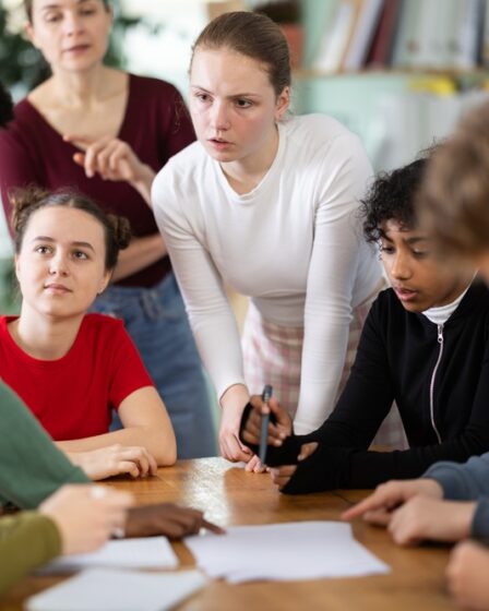 Alunos envolvidos discutindo e realizando uma atividade extensionista à mesa enquanto o professor observa seu progresso.