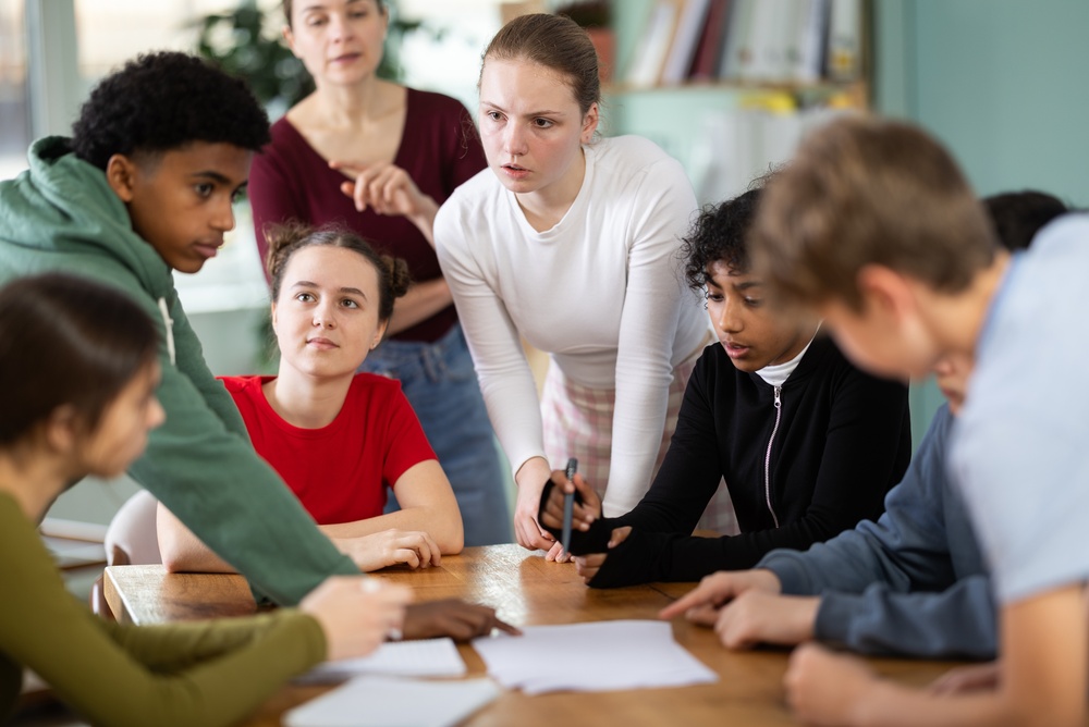 Alunos envolvidos discutindo e realizando uma atividade extensionista à mesa enquanto o professor observa seu progresso.