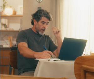 Homem sentado em frente ao notebook fazendo anotações durante estudo a distância.