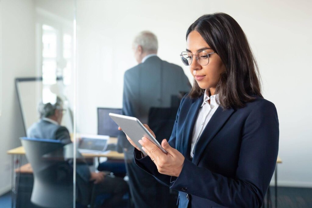 Mulher utilizando um tablet em um ambiente corporativo, com profissionais ao fundo em uma sala de reuniões.