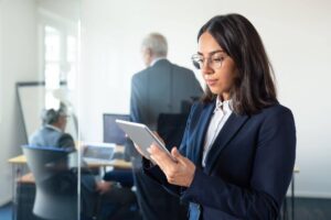 Mulher utilizando um tablet em um ambiente corporativo, com profissionais ao fundo em uma sala de reuniões.
