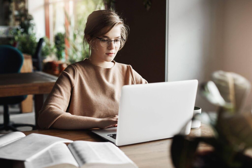 Mulher utilizando um notebook para estudar, com livros abertos e uma caneca sobre a mesa em um ambiente de estudo.
