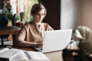 Mulher utilizando um notebook para estudar, com livros abertos e uma caneca sobre a mesa em um ambiente de estudo.