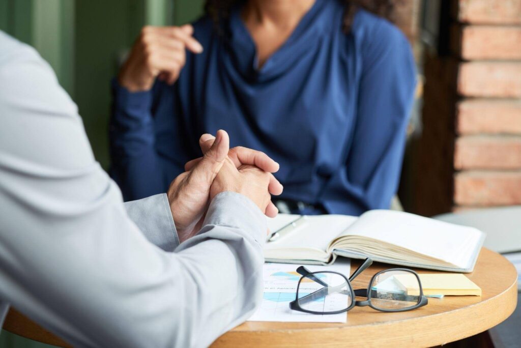 Pessoa sentada com as mãos entrelaçadas conversa com um terapeuta que faz anotações durante uma sessão de atendimento psicológico.