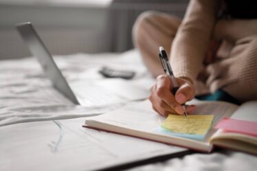 Pessoa escreve anotações em um caderno com apoio de um notebook e notas adesivas, organizando os estudos por meio de técnicas de memorização.