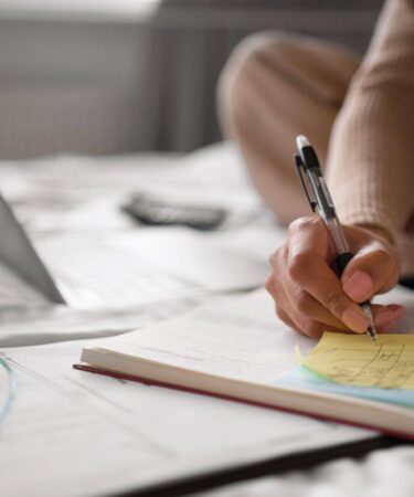 Pessoa escreve anotações em um caderno com apoio de um notebook e notas adesivas, organizando os estudos por meio de técnicas de memorização.