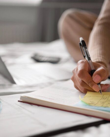 Pessoa escreve anotações em um caderno com apoio de um notebook e notas adesivas, organizando os estudos por meio de técnicas de memorização.