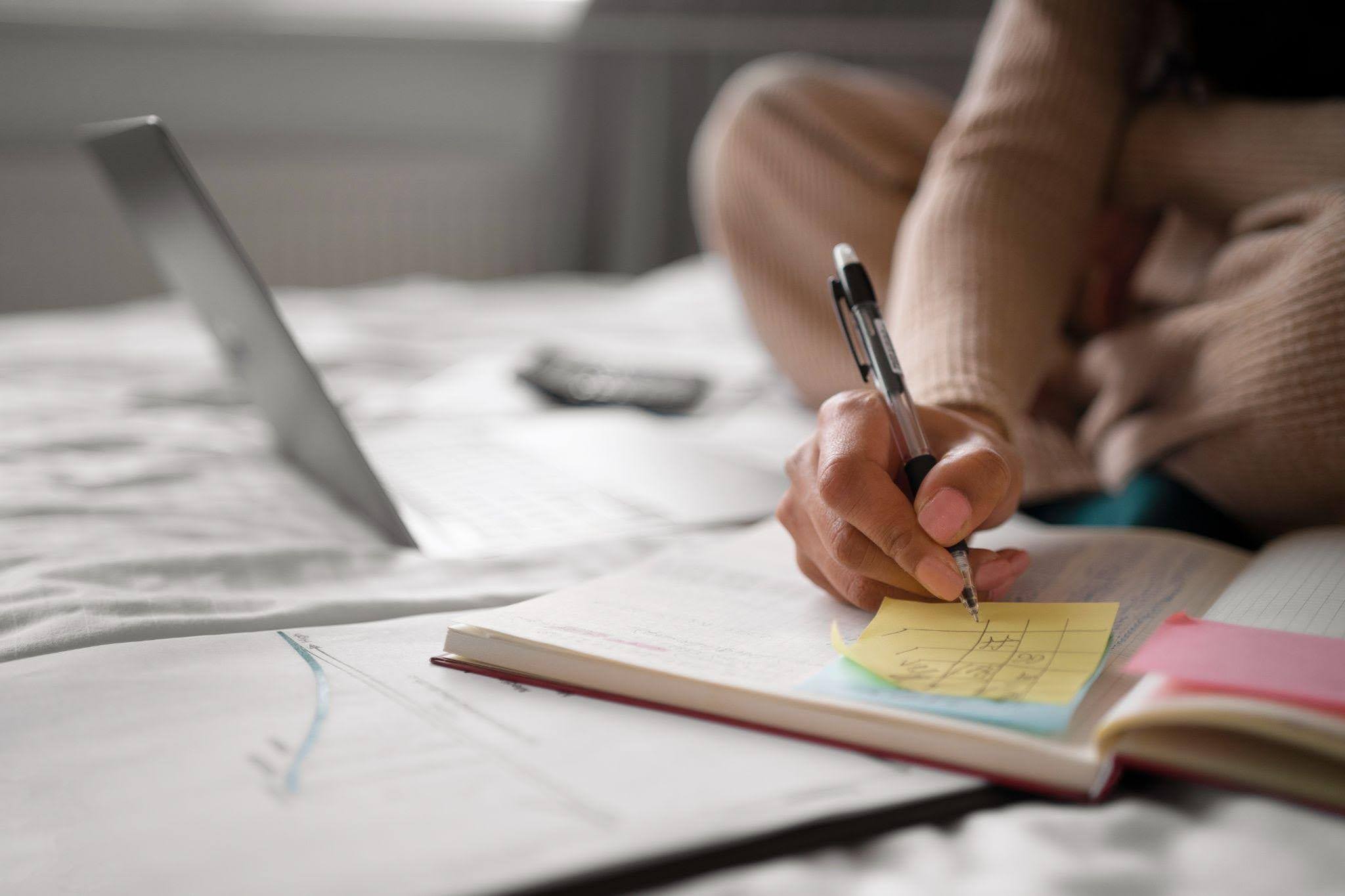 Pessoa escreve anotações em um caderno com apoio de um notebook e notas adesivas, organizando os estudos por meio de técnicas de memorização.