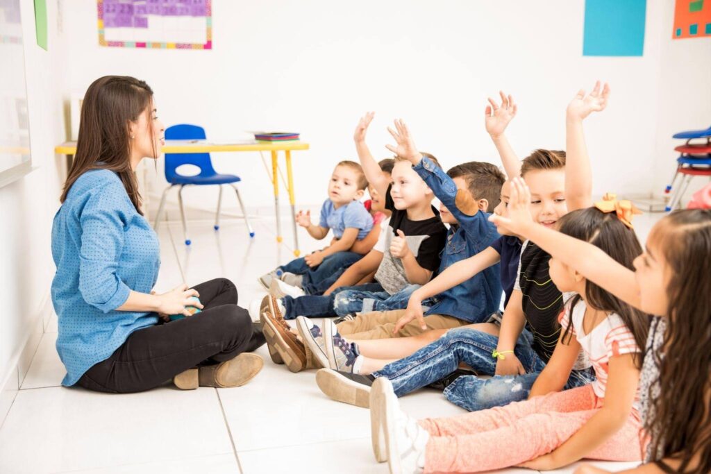 A professora sentada no chão conduz uma atividade com crianças da educação infantil, ilustrando metodologias de ensino abordadas na Licenciatura em Pedagogia.