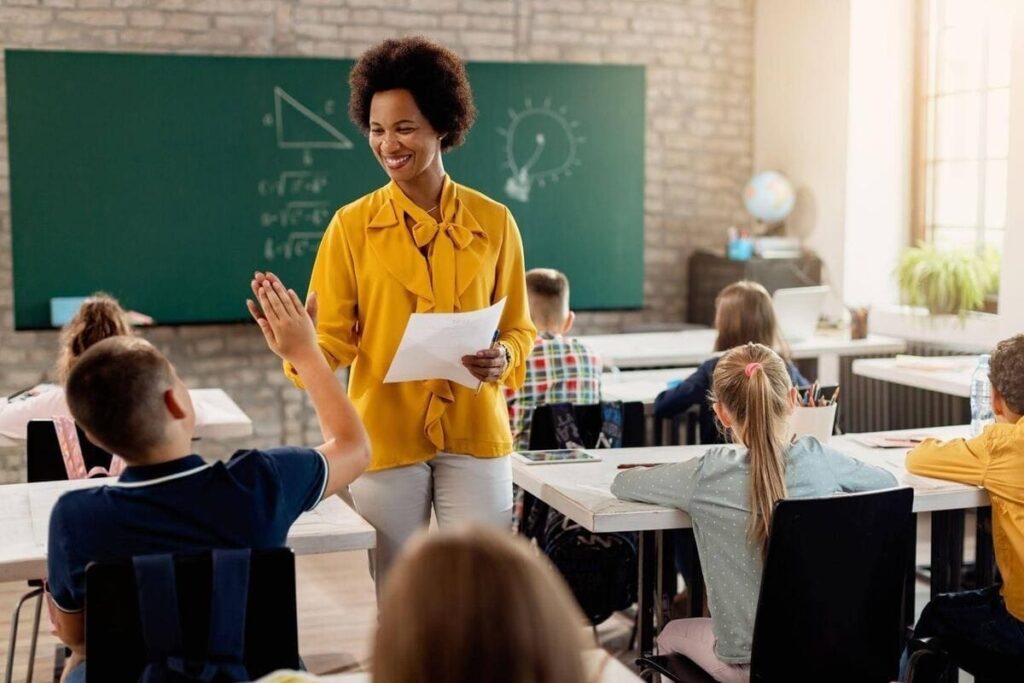 Professora interagindo com um aluno que levanta a mão durante uma aula em sala de aula. Cena representa a prática docente desenvolvida na Licenciatura em Pedagogia.