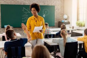 Professora interagindo com um aluno que levanta a mão durante uma aula em sala de aula. Cena representa a prática docente desenvolvida na Licenciatura em Pedagogia.