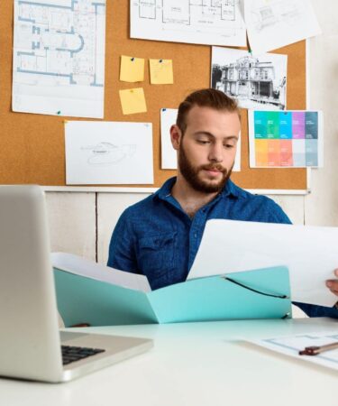 Homem usando óculos, analisando um documento enquanto trabalha em uma mesa com papéis, notebook, celular, e marcadores coloridos ao fundo.