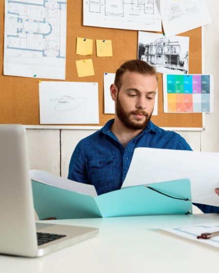 Homem usando óculos, analisando um documento enquanto trabalha em uma mesa com papéis, notebook, celular, e marcadores coloridos ao fundo.