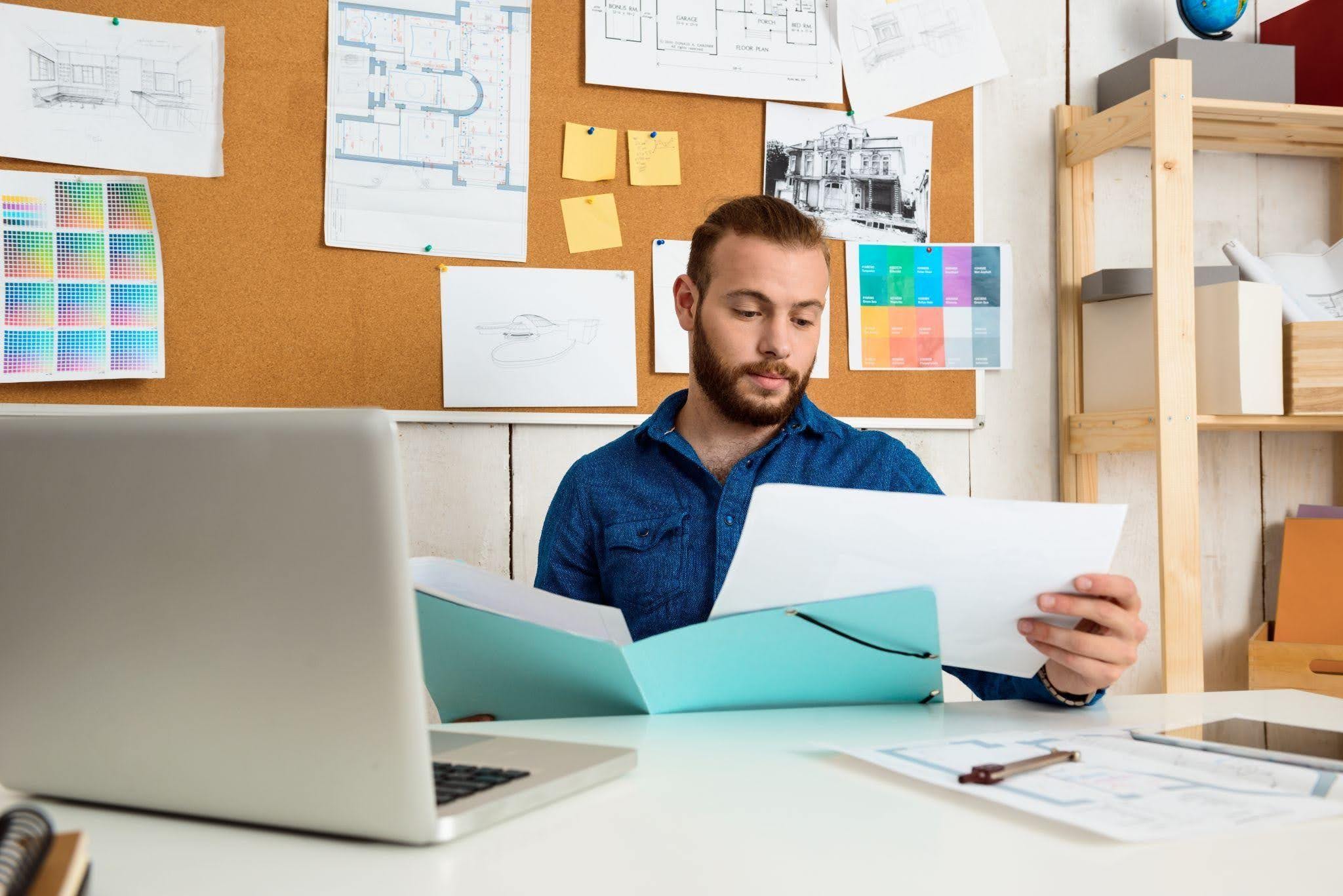 Homem usando óculos, analisando um documento enquanto trabalha em uma mesa com papéis, notebook, celular, e marcadores coloridos ao fundo.