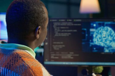 Homem trabalhando em computador com códigos de programação e imagem de inteligência artificial na tela.