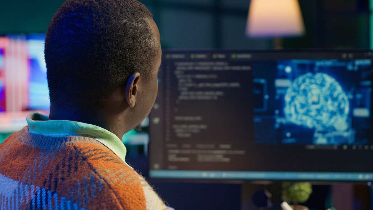 Homem trabalhando em computador com códigos de programação e imagem de inteligência artificial na tela.