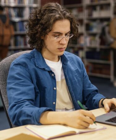 Pessoa estudando em uma biblioteca, fazendo anotações em um caderno enquanto utiliza um notebook.