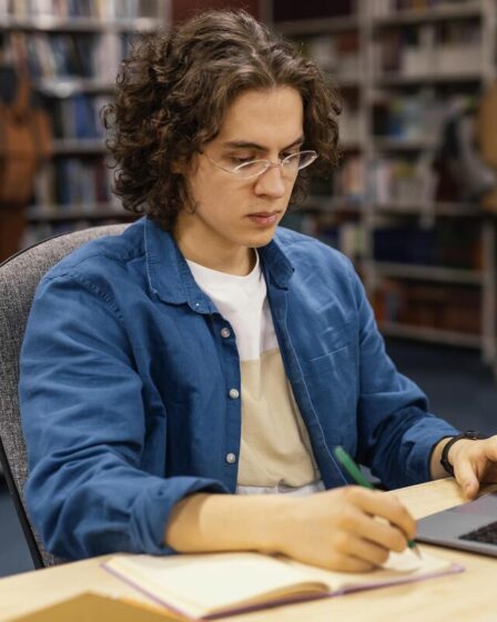 Pessoa estudando em uma biblioteca, fazendo anotações em um caderno enquanto utiliza um notebook.