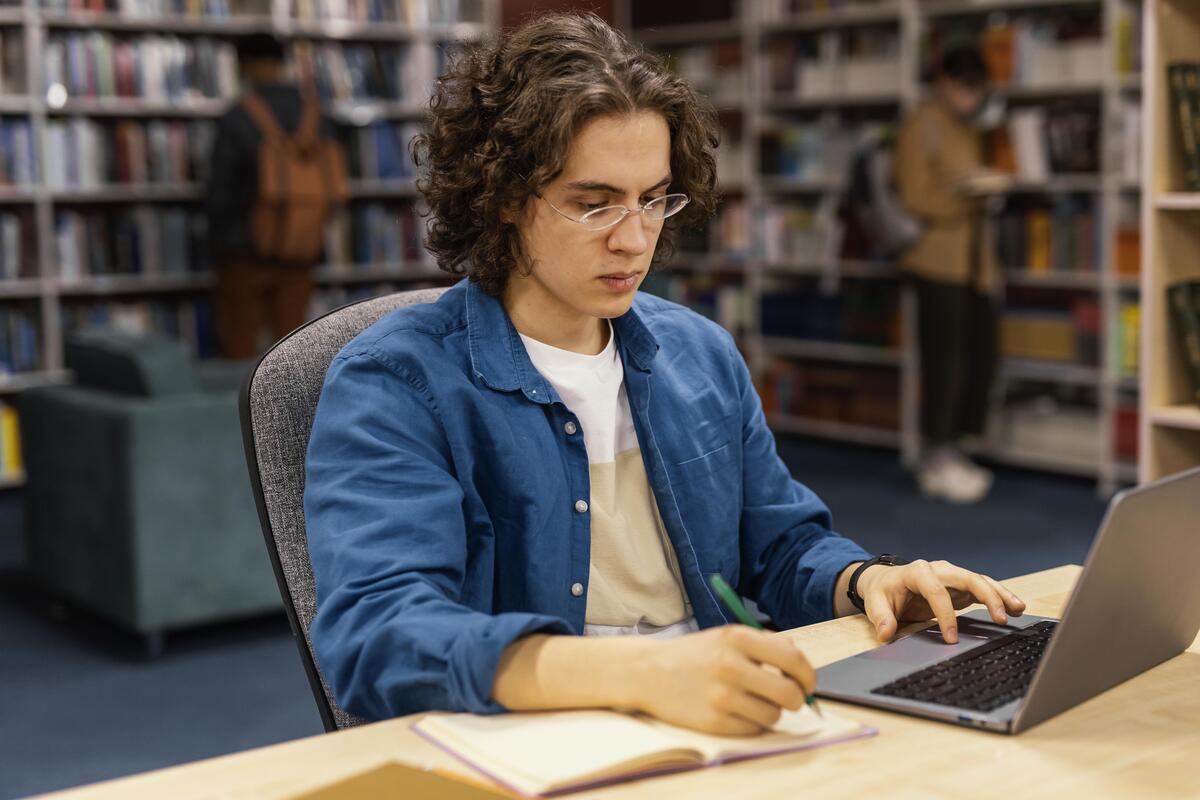 Pessoa estudando em uma biblioteca, fazendo anotações em um caderno enquanto utiliza um notebook.