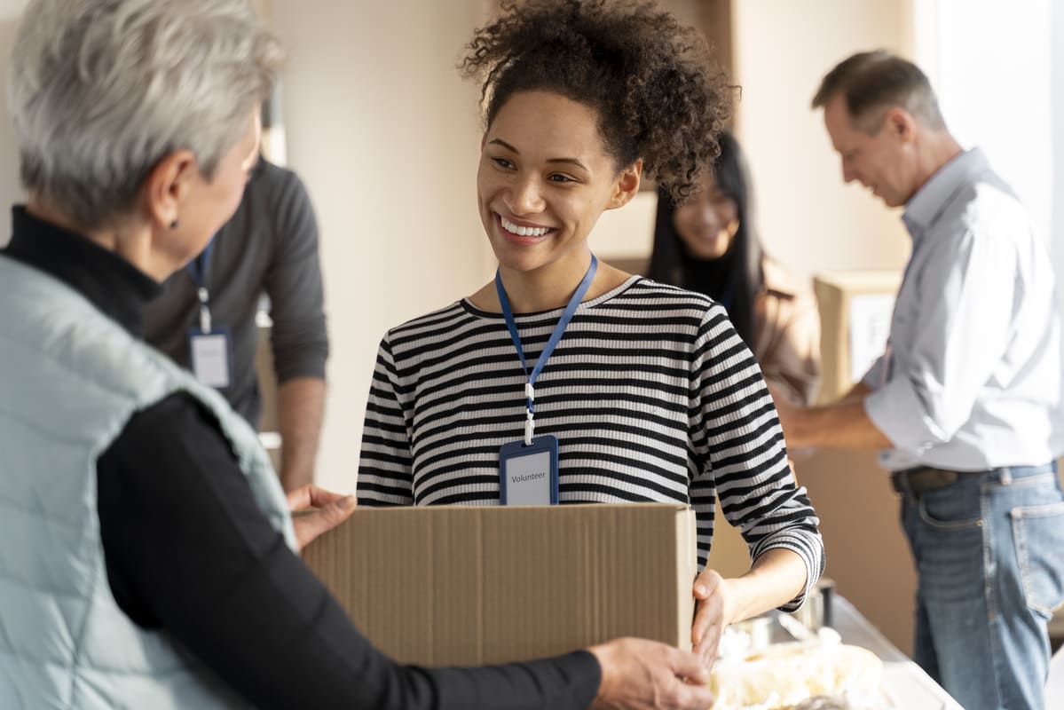 Voluntária sorridente entrega uma caixa de doações a outra pessoa, representando o que é Serviço Social: solidariedade e apoio à comunidade.