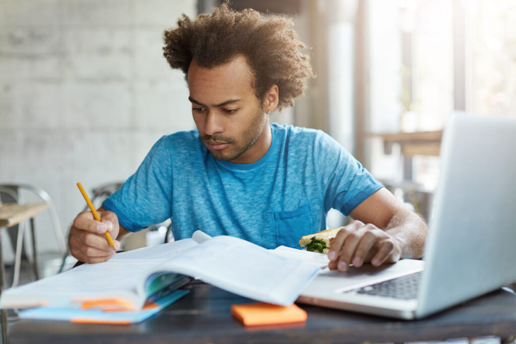 Homem estudando para uma prova, escrevendo anotações em um caderno enquanto utiliza um notebook.