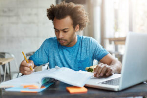 Homem estudando para uma prova, escrevendo anotações em um caderno enquanto utiliza um notebook.