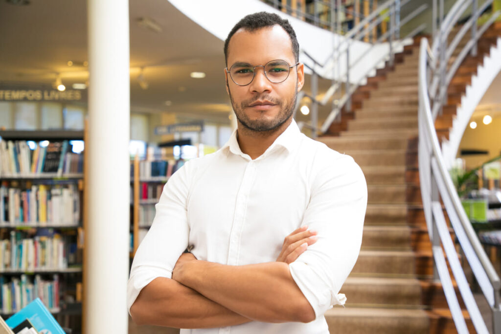 Professor em pé com os braços cruzados dentro de uma biblioteca, representando o estudo acadêmico sobre Sociologia e a análise das relações sociais.