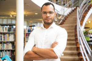 Professor em pé com os braços cruzados dentro de uma biblioteca, representando o estudo acadêmico sobre Sociologia e a análise das relações sociais.