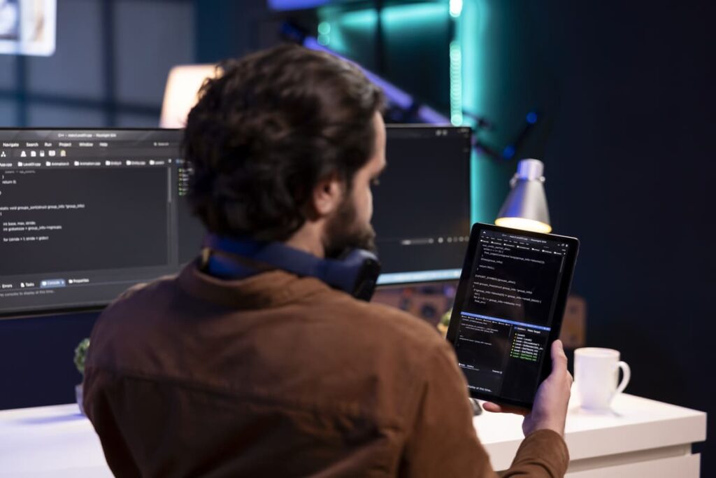 Profissional de tecnologia analisando código em um tablet diante de múltiplos monitores, ilustrando processos de Engenharia de Software para Dispositivos Móveis.