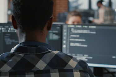 Desenvolvedor trabalhando em frente a monitores com código em ambiente corporativo, demonstrando atividades de Engenharia de Software para Dispositivos Móveis.