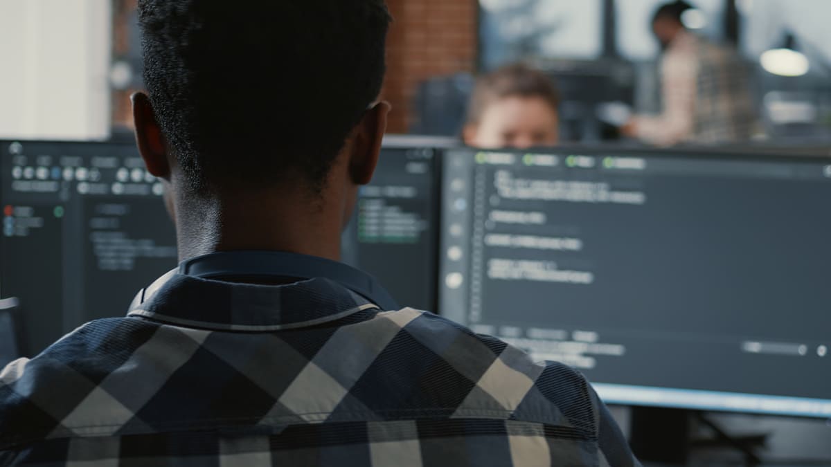Desenvolvedor trabalhando em frente a monitores com código em ambiente corporativo, demonstrando atividades de Engenharia de Software para Dispositivos Móveis.