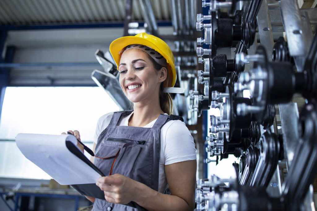 Profissional da Engenharia de Fabricação Mecânica utilizando capacete de segurança e prancheta, fazendo inspeção e controle de qualidade em equipamentos industriais.