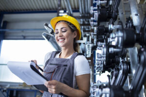 Profissional da Engenharia de Fabricação Mecânica utilizando capacete de segurança e prancheta, fazendo inspeção e controle de qualidade em equipamentos industriais.