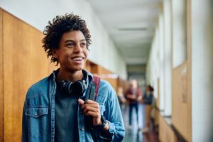 Jovem estudante do ensino médio sorri enquanto olha para alto. Atrás dele, estudantes conversam em um corredor de uma escola.