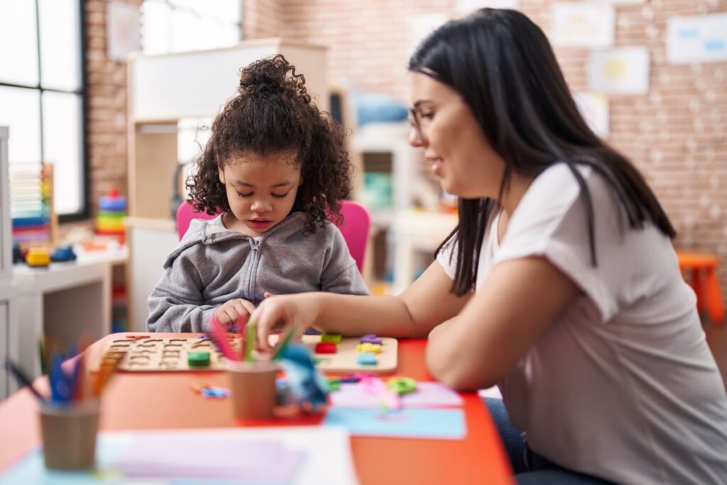 Professora acompanhando uma criança durante atividade educativa com peças coloridas e jogos de aprendizagem.
