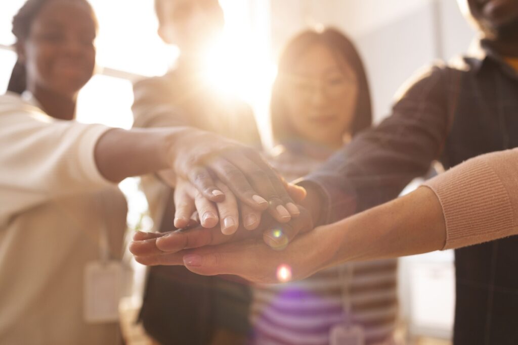Grupo diverso de pessoas coloca as mãos umas sobre as outras em um momento de união, representando o que é Serviço Social: cooperação e trabalho em equipe.