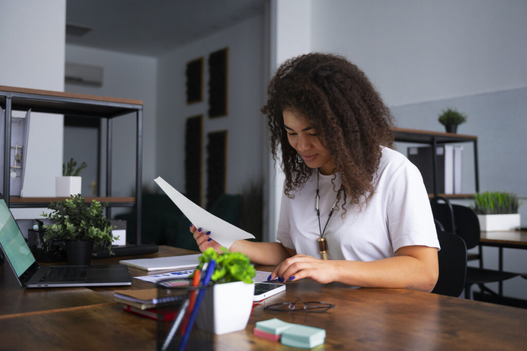 Mulher estudando em um escritório, analisando um documento enquanto trabalha com materiais de estudo e um notebook.