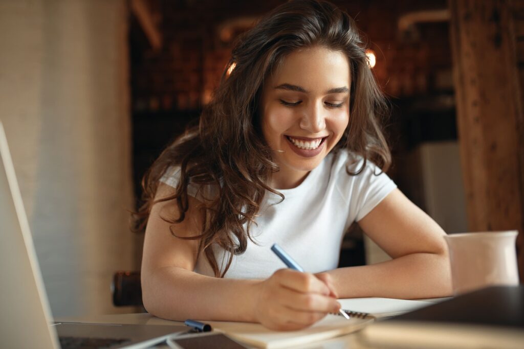 Estudante sorridente faz anotações enquanto estuda com um notebook.