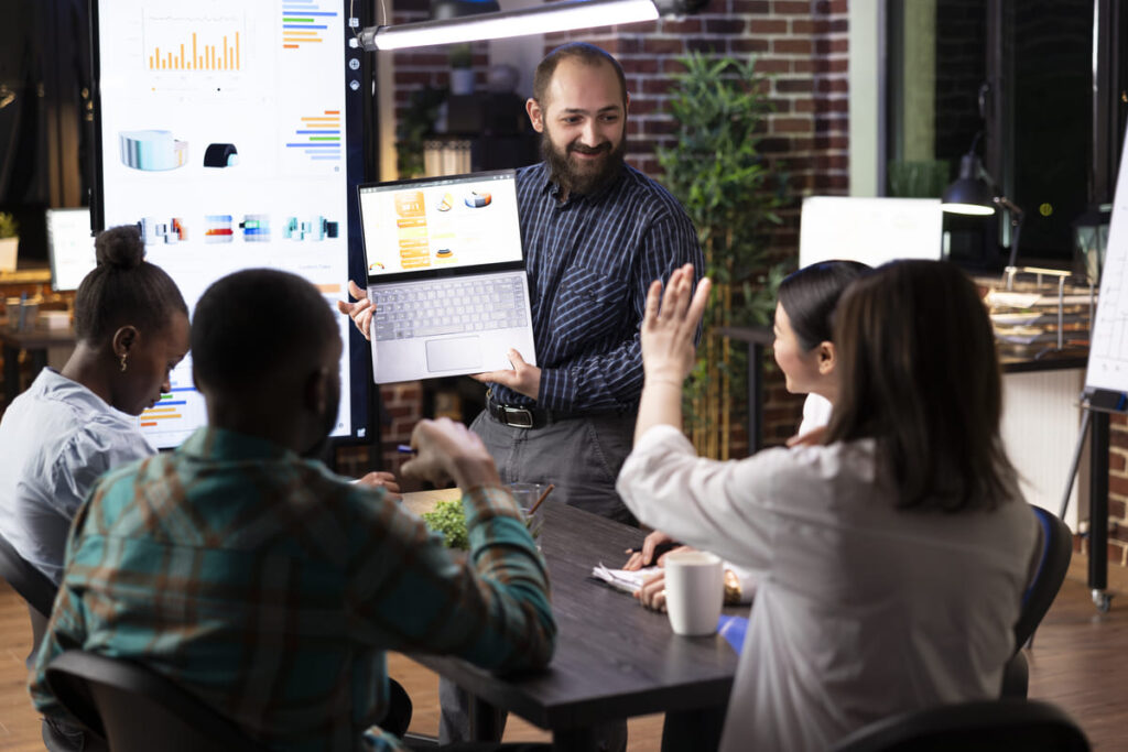 Equipe de profissionais em uma reunião de trabalho. Um homem apresenta dados em um notebook enquanto os demais participantes observam e interagem à mesa. 