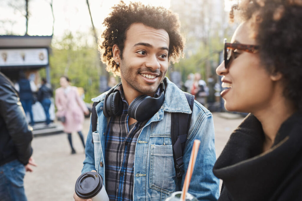 Jovem sorrindo, jaqueta jeans e fones de ouvido no pescoço, conversando com uma mulher em um parque enquanto segura um copo de café.