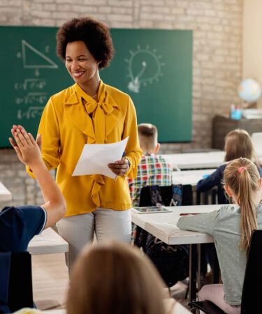 Professora sorrindo em sala de aula enquanto interage com os alunos, ilustrando práticas de ensino e formação em pós-graduação em Educação.