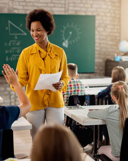 Professora sorrindo em sala de aula enquanto interage com os alunos, ilustrando práticas de ensino e formação em pós-graduação em Educação.