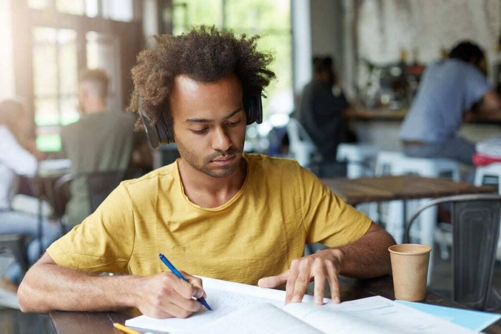 Jovem sentado à mesa, usando fones de ouvido e escrevendo em um caderno.