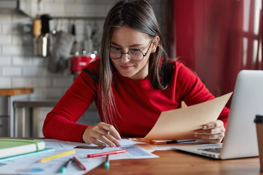 Mulher usando óculos e blusa vermelha, sentada à mesa com documentos e um lápis, analisando uma folha ao lado de um notebook aberto.