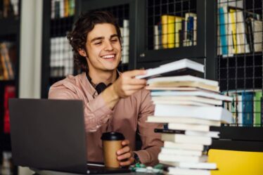 Estudante sorridente segura livros ao lado de um notebook durante os estudos.